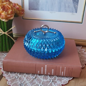 May include: A vintage, faceted, turquoise-blue glass trinket box with a silver-tone handle. The box is round and sits atop a book titled "The Modern Handbook of Humor".