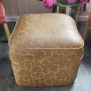 May include: A vintage square ottoman with a gold and brown floral pattern. The top and sides are upholstered with a textured material. The ottoman has a gold trim and sits on a grey floor. Decorative flowers are in the background.