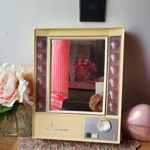 Puede incluir: Un espejo Clairol True-Light vintage con un marco color crema y un espejo rectangular. El espejo tiene un selector de luz y una serie de luces redondas teñidas de rosa a cada lado. El texto "Clairol True-Light Mirror" está impreso en la parte delantera.