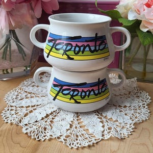 May include: Two white ceramic soup bowls with handles. Both bowls have a colorful rainbow stripe design with the word "Toronto" written in black. The bowls are stacked on top of each other.