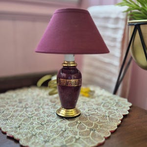 May include: A miniature table lamp featuring a deep maroon fabric lampshade and a matching maroon ceramic base. The base is accented with a gold-coloured band featuring intricate black patterns and a polished gold-coloured circular pedestal base.