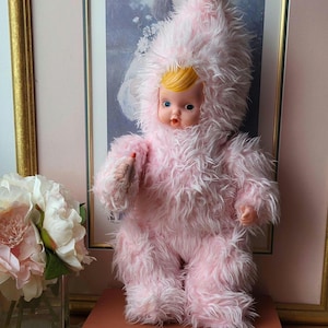 Puede incluir: Una muñeca vintage con un atuendo rosa y peludo con capucha. La muñeca tiene una cara de plástico con ojos azules y cabello rubio. La muñeca está de pie sobre un libro. El fondo incluye un cuadro enmarcado y un jarrón con flores.