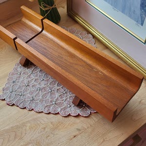 Puede incluir: Una bandeja de madera para servir con un diseño largo en forma de artesa. La bandeja está hecha de madera de tonos cálidos y se eleva sobre pequeños soportes de madera. La bandeja se encuentra sobre un mantel individual decorativo con cuentas.
