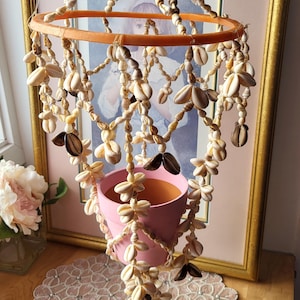 Könnte beinhalten: Ein hängender Pflanztopf mit einem rosa Topf, der von einem dekorativen Muschel-Mobile abgehängt ist. Das Mobile hat einen orangefarbenen Ring und Stränge aus verschiedenen Muscheln. Der Pflanztopf steht auf einem Perlen-Platzset, mit einem gerahmten Bild im Hintergrund.