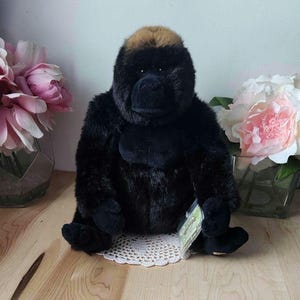 May include: A black and brown plush gorilla toy sitting on a white crocheted doily. The gorilla has a tag attached to its leg.