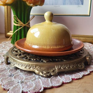 May include: A yellow and brown ceramic butter dish with a lid, on a decorative gold-coloured metal trivet. The trivet rests on a pink beaded placemat. A bouquet of orange and yellow flowers is in the background.