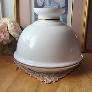 May include: A white, glossy, glass lamp shade with a gold trim. The shade is dome-shaped and sits on a decorative, lace-edged mat. A framed portrait and pink flowers are in the background.