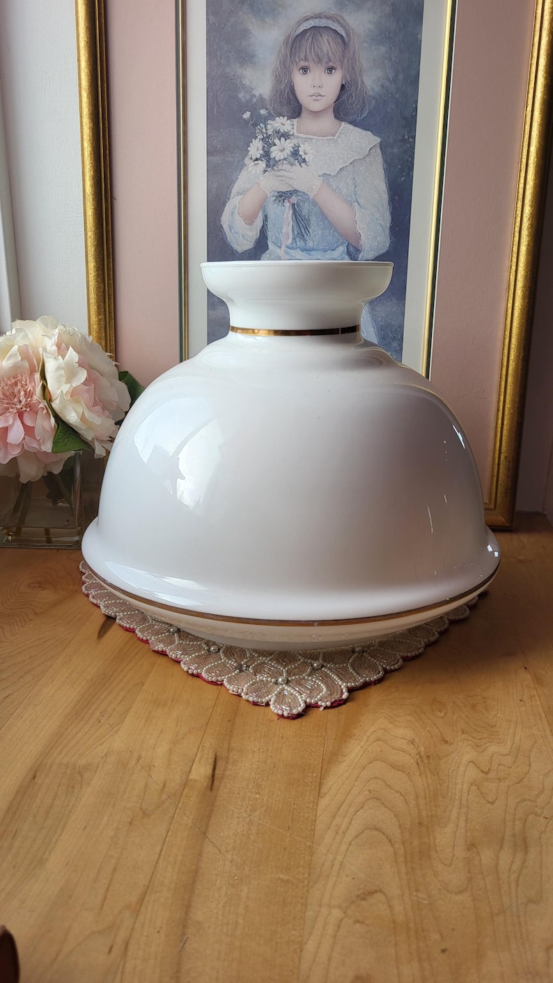 May include: A white, glossy, glass lamp shade with a gold trim. The shade is dome-shaped and sits on a decorative, lace-edged mat. A framed portrait and pink flowers are in the background.