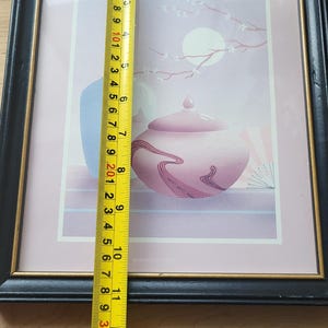 Op de afbeelding: Een ingelijste print van een Japans ge&iuml;nspireerd stilleven met een roze en paarse theepot, een blauwe vaas en een tak met witte bloemen op een lichtroze achtergrond. De print is ingelijst in een zwart frame met een gouden binnenrand.