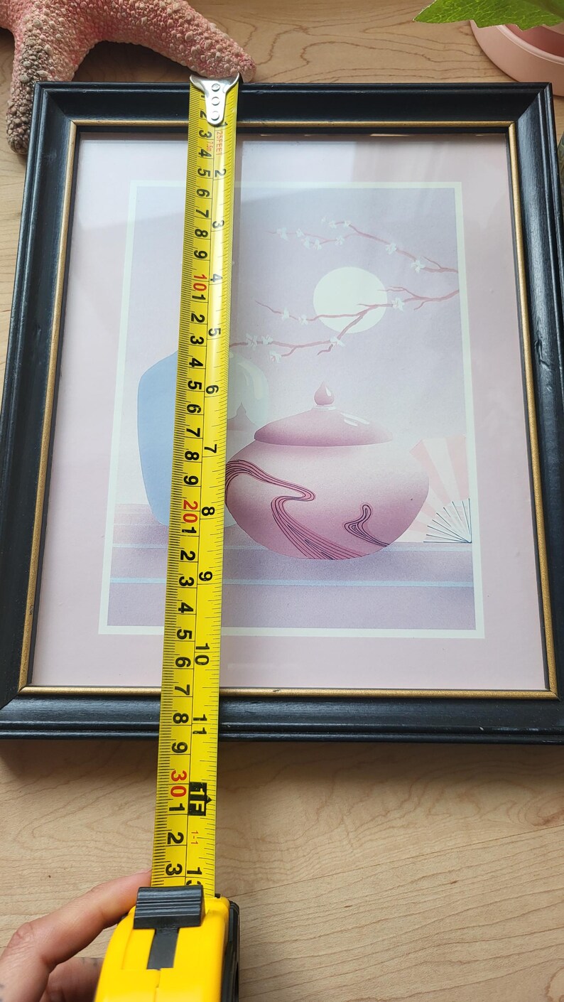 Op de afbeelding: Een ingelijste print van een Japans ge&iuml;nspireerd stilleven met een roze en paarse theepot, een blauwe vaas en een tak met witte bloemen op een lichtroze achtergrond. De print is ingelijst in een zwart frame met een gouden binnenrand.