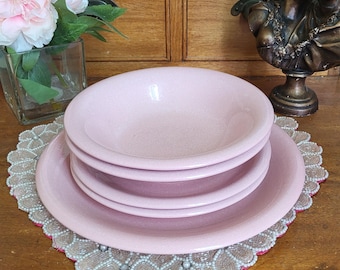 May include: A stack of pink ceramic bowls and plates on a decorative doily with a beaded edge. The dishes are set on a wooden surface, with a vase of pink flowers and a bronze bust in the background.