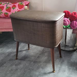 May include: A vintage sewing box with a brown, textured exterior and a padded lid. The box is rectangular with rounded corners and stands on four slender, dark wooden legs with brass-colored tips. The box is a classic piece of home decor.