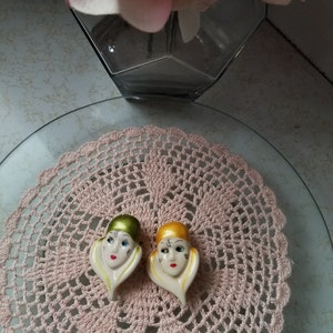 K&ouml;nnte beinhalten: Zwei Keramikfiguren von Frauenk&ouml;pfen mit stilisierten Gesichtsz&uuml;gen. Die Figuren tragen H&uuml;te, einer ist gr&uuml;n und der andere ist gelb. Die Figuren stehen auf einer rosa H&auml;keldeckchen.