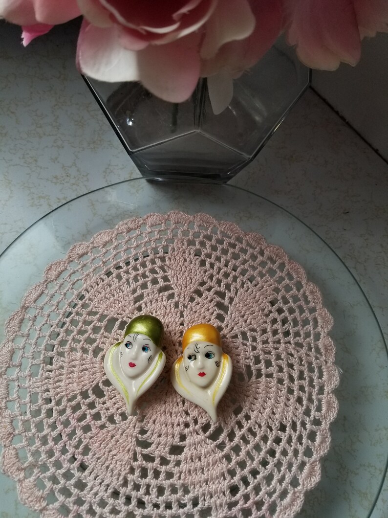 K&ouml;nnte beinhalten: Zwei Keramikfiguren von Frauenk&ouml;pfen mit stilisierten Gesichtsz&uuml;gen. Die Figuren tragen H&uuml;te, einer ist gr&uuml;n und der andere ist gelb. Die Figuren stehen auf einer rosa H&auml;keldeckchen.