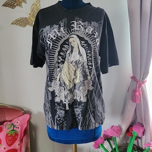 May include: Black t-shirt featuring a graphic of a religious figure with hands clasped in prayer, surrounded by roses and text. The design is in shades of gray and white. The shirt is displayed on a mannequin.