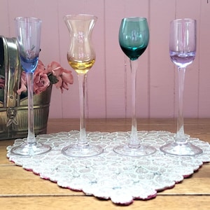 Puede incluir: Cuatro copas de licor de colores con tallos largos. Las copas tienen cuencos teñidos de azul, amarillo, verde y morado. Están dispuestas sobre un tapete de encaje blanco con ribete rosa, sobre una superficie de madera.