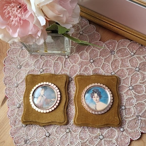 Puede incluir: Dos miniaturas de retratos ovalados antiguos en marcos dorados ornamentados, sobre cojines cuadrados de terciopelo verde oliva. Los retratos representan figuras con vestimenta de época. La exhibición está sobre una alfombra con cuentas y motivos florales.