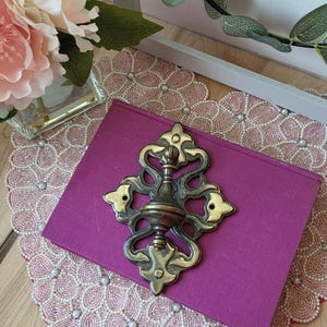 May include: An ornate brass drawer pull rests on a purple book. The image also features a beaded placemat, a floral arrangement, and a sign that says "bonheur".