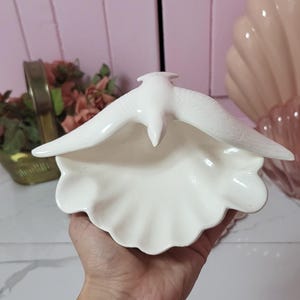 May include: A white ceramic dish in the shape of a seashell, with a bird figurine perched above. The bird's wings are outstretched, and the dish has a ruffled edge. The background is a soft pink.