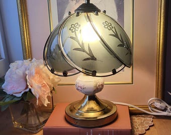 May include: A vintage table lamp with a floral design on the shade and base. The lamp has a brass base and a light green shade with black floral accents. It sits on a book, next to a vase of flowers.