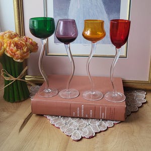 May include: Four coloured stemmed glasses with curved stems. The glasses are green, purple, orange, and red. They are arranged on a book titled "The Modern Handbook of Humor". A painting of a girl is in the background.