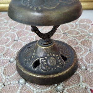 Puede incluir: Un quemador de incienso antiguo de color bronce con motivos florales. El quemador tiene una parte superior en forma de c&uacute;pula y una base circular con recortes decorativos. El dise&ntilde;o incluye un asa curva que conecta la parte superior e inferior. El quemador de incienso es un art&iacute;culo decorativo.