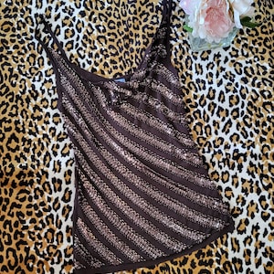 May include: A brown sequined vest top with diagonal stripes, set against a leopard print fabric. The top features thin straps and a ruffled neckline. A small bouquet of pink flowers in a glass vase is in the background.