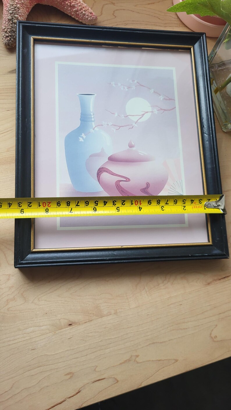 Op de afbeelding: Een ingelijste print van een stilleven met een blauwe vaas en een roze pot met deksel met een bloemenmotief op een lichtroze achtergrond.