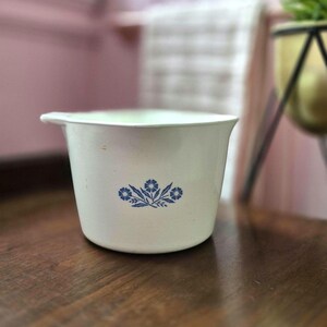 Puede incluir: Un cuenco mezclador de cerámica blanca con borde acampanado y un pequeño pico vertedor. El exterior presenta un patrón floral azul centrado que consiste en tres flores estilizadas con hojas, dispuestas en un diseño horizontal simétrico.