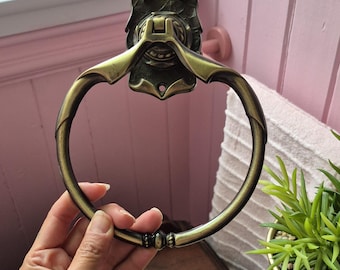 May include: A circular towel ring with an antique brass finish, featuring a decorative wall-mounting plate with two screw holes and a hinged mechanism at the top that allows the ring to pivot downward from its vertical position.