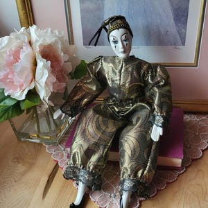 May include: A porcelain doll in a gold and black patterned harlequin costume, with a matching hat. The doll has a painted face with blue eyes and black accents. It is seated on a purple book.