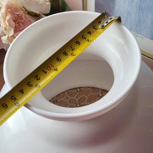 May include: A white ceramic vase with a wide opening, measured with a yellow tape measure. The tape measure shows a measurement of approximately 6 inches. The vase has a round base and a textured interior.