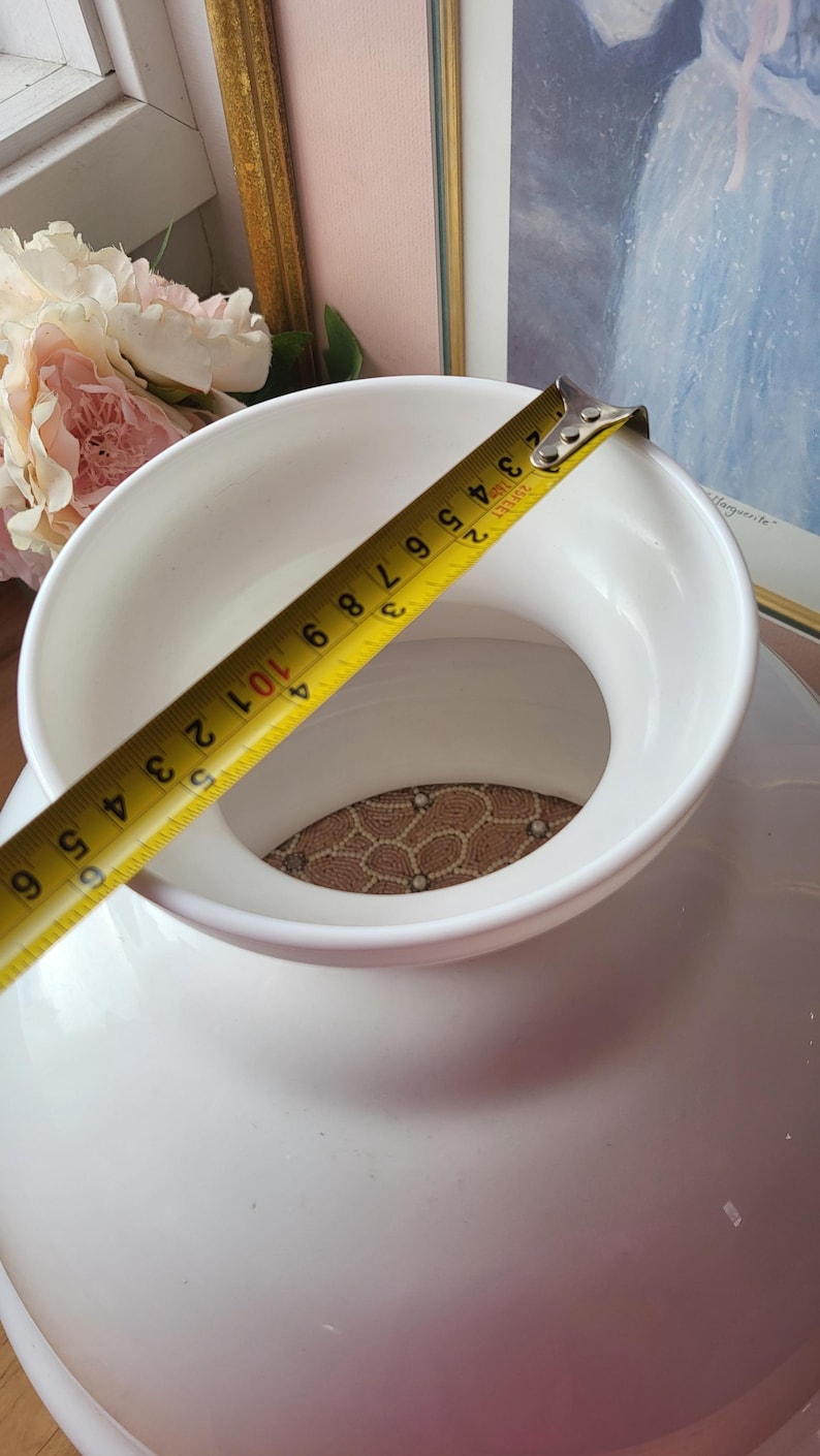 May include: A white ceramic vase with a wide opening, measured with a yellow tape measure. The tape measure shows a measurement of approximately 6 inches. The vase has a round base and a textured interior.