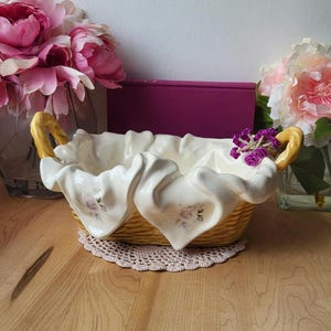May include: A ceramic basket shaped like a woven basket with white fabric draped over the sides. The basket is decorated with floral designs and has a yellow glaze. The basket is sitting on a crocheted doily.