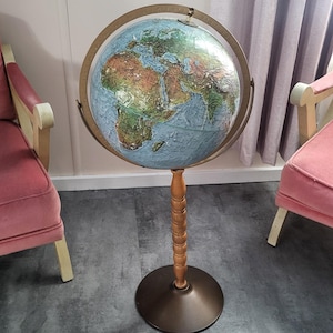 Może przedstawiać: Zabytkowy globus na stojaku. Globus przedstawia szczegółową mapę z niebieskimi oceanami i brązowymi oraz zielonymi lądami. Jest podparty drewnianą kolumną i ciemnobrązową podstawą. Globus otoczony jest brązowym pierścieniem południka.