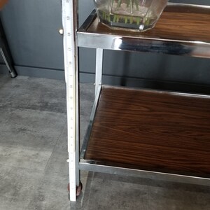 May include: A chrome bar cart with two wooden shelves. The cart has a single wheel on the bottom left side and is 21 inches tall. The top shelf has a glass vase with pink flowers.