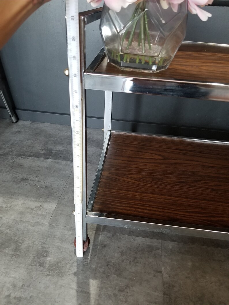May include: A chrome bar cart with two wooden shelves. The cart has a single wheel on the bottom left side and is 21 inches tall. The top shelf has a glass vase with pink flowers.