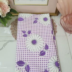Puede incluir: Tela doblada con un estampado floral de margaritas blancas y moradas sobre un fondo a cuadros morado. Las margaritas tienen pétalos blancos y centros morados. La tela está doblada cuidadosamente.