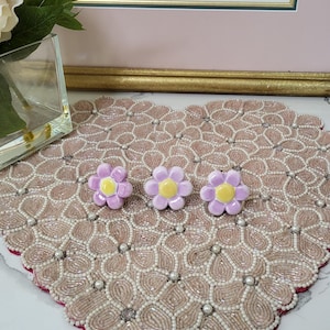 May include: Three ceramic flower-shaped knobs with lavender petals and yellow centres are displayed on a beaded placemat. The placemat is a light pink colour with a floral pattern and pearl accents. A framed artwork is in the background.