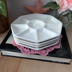 May include: A set of four white ceramic octagonal serving dishes with six individual compartments. The dishes are stacked on top of each other and are sitting on a pink doily.