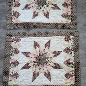 Pode incluir: Duas toalhas de mesa acolchoadas com um design de estrela. O centro de cada toalha de mesa apresenta um padrão de estrela em tons de bordô e creme, rodeado por padrões florais e geométricos. Uma borda marrom estampada emoldura o design.