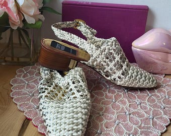 以下が含まれることがあります： ヘビ柄の織りオープントゥサンダル。ヒールは茶色で、ストラップにはゴールドのバックルが付いています。靴はピンク色のビーズのプレースマットの上に展示されています。