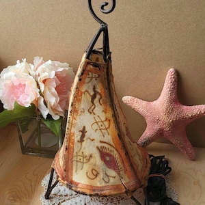 Op de afbeelding: Een tafellamp met een unieke, handgeschilderde leren kap met een geometrisch patroon en een rood oogontwerp. De lamp heeft een zwarte metalen voet en een gekrulde metalen bovenkant.