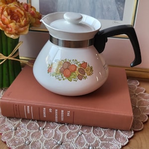 Könnte beinhalten: Weiße Keramik-Teekanne mit schwarzem Griff und Deckel. Die Teekanne hat ein dekoratives Band mit einem Fruchtdesign. Sie steht auf einem korallenfarbenen Buch und einer Perlen-Tischdecke, mit einem gerahmten Bild im Hintergrund.