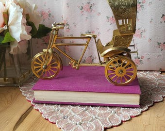 May include: A gold-coloured, decorative rickshaw model with intricate detailing, resting on a purple book. The rickshaw has a canopy and spoked wheels. The background features floral wallpaper and a decorative doily.