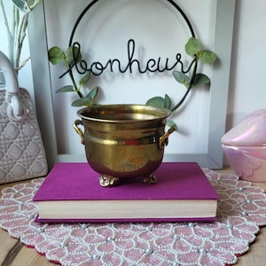 May include: A small, round, polished brass pot with two side handles and three decorative feet, resting on a thick, magenta-coloured hardcover book. The pot has a wide opening and a smooth, metallic finish that reflects light across its surface.