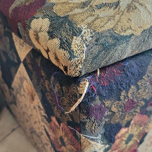 May include: Close-up of a floral patterned fabric ottoman with a frayed seam. The fabric is a mix of dark blue, burgundy, beige, and gold.