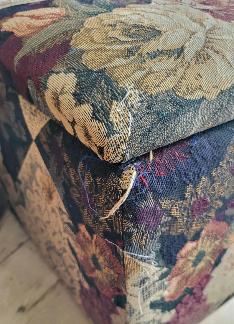 May include: Close-up of a floral patterned fabric ottoman with a frayed seam. The fabric is a mix of dark blue, burgundy, beige, and gold.