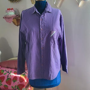 Peut inclure: Une chemise boutonnée violette à manches longues et à rayures verticales, présentée sur un mannequin. La chemise a une poche poitrine avec un petit motif coloré. Le mannequin est sur un support en laiton, et l'arrière-plan comprend des décorations murales.