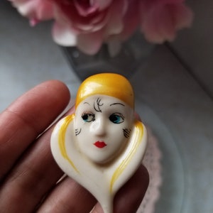 K&ouml;nnte beinhalten: Eine wei&szlig;e Keramikbrosche mit einem bemalten Gesicht und gelben Haaren. Das Gesicht hat schwarze Augen, einen roten Mund und schwarze Markierungen.